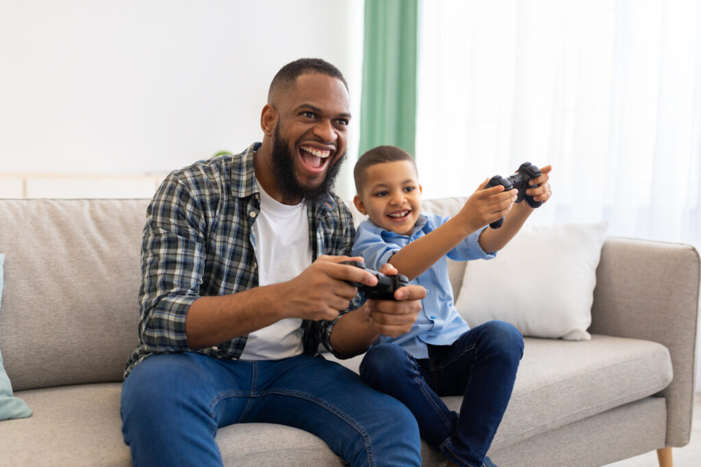 Joyful African Father And His Kid Boy Playing Videogames Bonding And Spending Time Together Having Fun On Weekend At Home. New Video Game Advertisement Concept